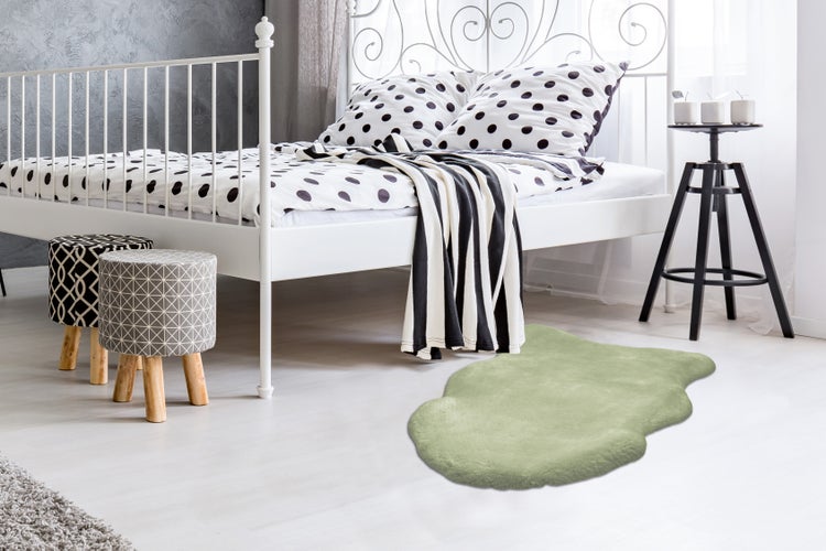 Světlá ložnice s bílou kovovou postelí a zeleným kobercem z umělé kožešiny na světlé podlaze. Vedle postele stojí dvě vzorované stoličky.