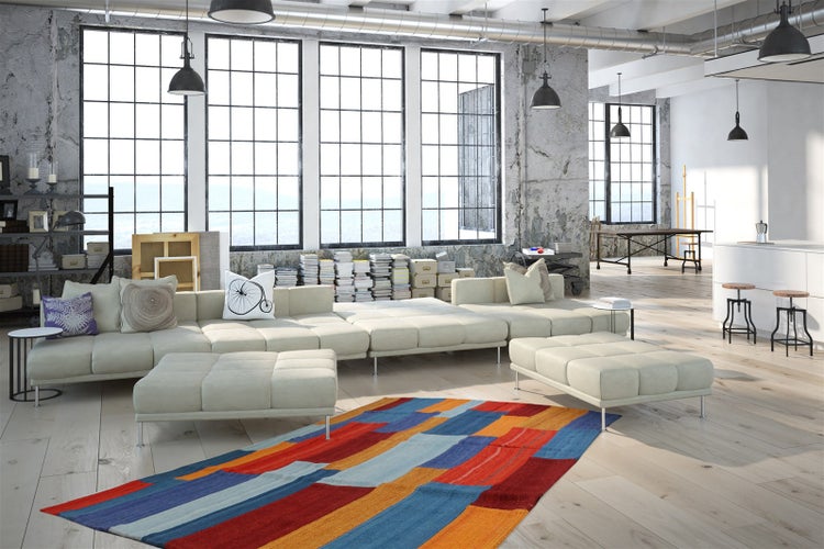 Industriální loft s velkou modulární pohovkou, barevným kobercem, světlou dřevěnou podlahou a betonovými stěnami.