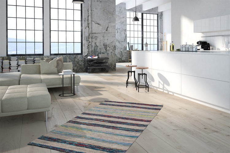 Moderní loft v industriálním stylu se světlou dřevěnou podlahou, pruhovaným kobercem, světlou modulární pohovkou, velkými okny a bílým ostrůvkem.
