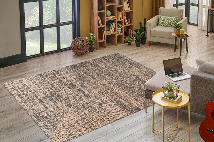 Obývací pokoj s kobercem s leopardím vzorem na světlé dřevěné podlaze, velkým oknem, dřevěnou knihovnou, křeslem a pokojovými rostlinami.