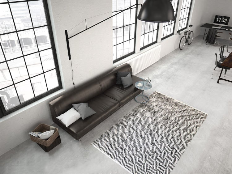 Moderní industriální loft s tmavou koženou pohovkou, geometrickým kobercem, velkými okny a černou nástěnnou lampou na betonové podlaze.