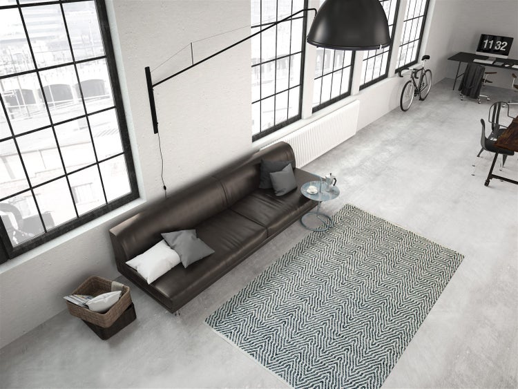 Moderní obývací prostor v industriálním stylu s hnědou koženou pohovkou, kobercem s geometrickým vzorem na betonové podlaze a velkými okny.