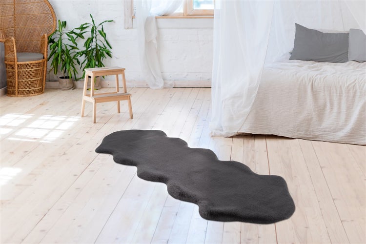 Šedý koberec z umělé kožešiny ve tvaru jehněčí kůže na světlé dřevěné podlaze ve světlé ložnici s postelí a rostlinami.