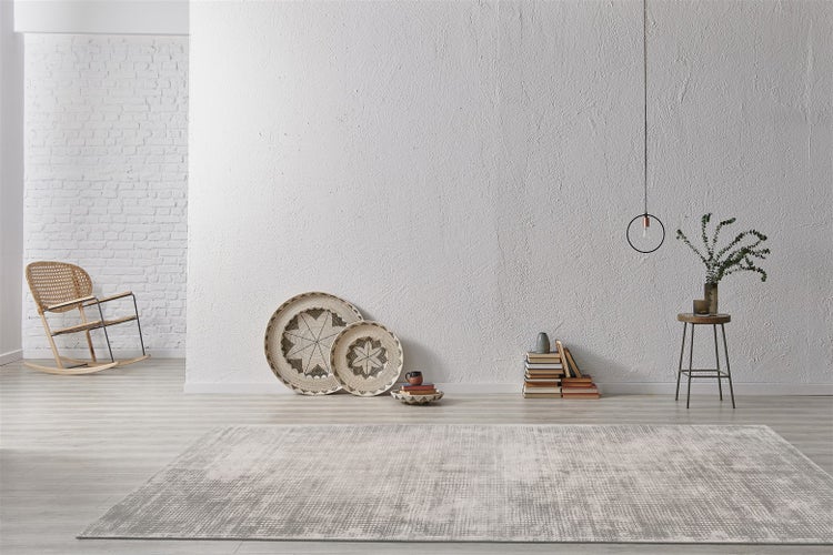 Moderní obývací pokoj s šedo-bílým vzorovaným kobercem na světlé podlaze, strukturovanou stěnou, proutěným křeslem a minimalistickou dekorací.