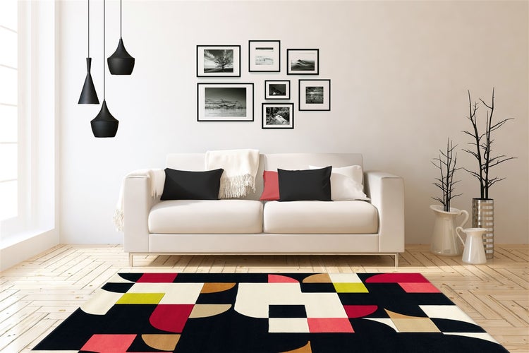 Moderní obývací pokoj s bílou pohovkou, barevným geometrickým kobercem, galerií obrazů a černými závěsnými svítidly na světlé podlaze.