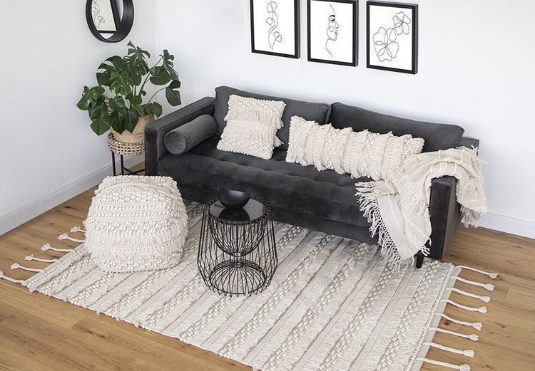 Moderní obývací pokoj s tmavě šedou pohovkou, krémovými makramé polštáři, ladícím taburetem a strukturovaným kobercem na dřevěné podlaze.