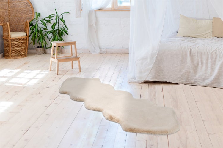 Dlouhý béžový koberec z umělé kožešiny s vlnitými okraji na světlé dřevěné podlaze ve světlé místnosti s postelí a proutěným křeslem.