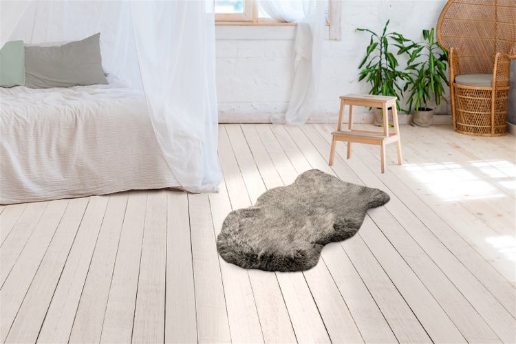 Koberec z umělé kožešiny ve vzhledu beránka na dřevěné podlaze ve světlé ložnici s postelí, dřevěnou stoličkou a rostlinami.