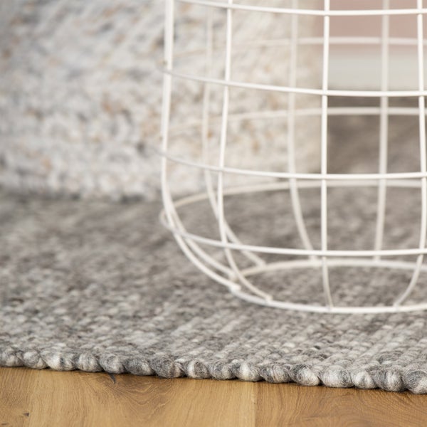 Detailní záběr na šedobílý melírovaný ručně vázaný vlněný koberec s hrubou strukturou na dřevěné podlaze vedle bílého drátěného koše.