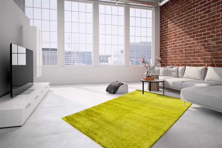 Moderní obývací pokoj v industriálním stylu s cihlovou stěnou, bílou pohovkou a jasně zeleným kobercem s vysokým vlasem na šedé podlaze.