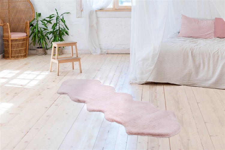 Růžový běhoun z umělé kožešiny nepravidelného tvaru na světlé dřevěné podlaze v ložnici s postelí a proutěným křeslem.