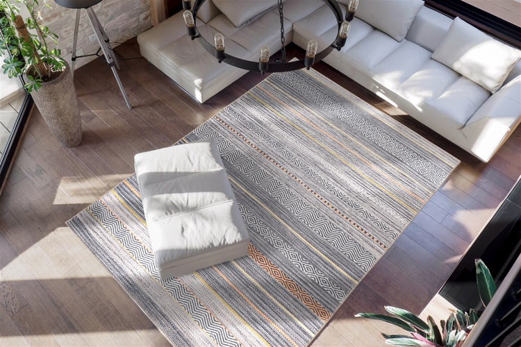 Moderní obývací pokoj s velkým vzorovaným kobercem v šedé a žluté barvě na tmavé dřevěné podlaze. Bílá kožená pohovka, taburet a světlý prostor.