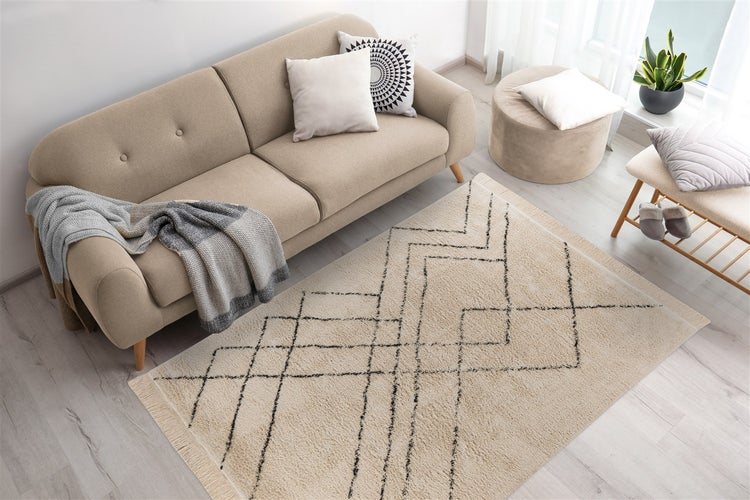 Světlý obývací pokoj s béžovou pohovkou, kobercem s černým geometrickým vzorem, pufem a dřevěnou lavicí na světlé dřevěné podlaze.