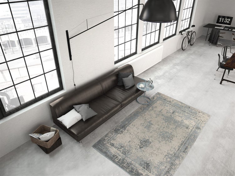 Prostorný industriální loft s velkými okny, šedo-béžovým kobercem ve vintage stylu na betonové podlaze a hnědou koženou pohovkou.