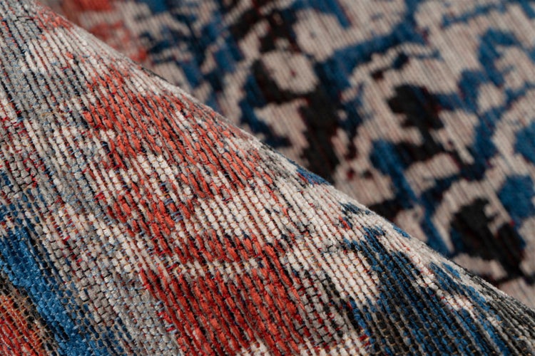 Detailní záběr modro-červeného tkaného koberce s vintage vzorem a výraznou texturou, jeden roh je přehnutý.