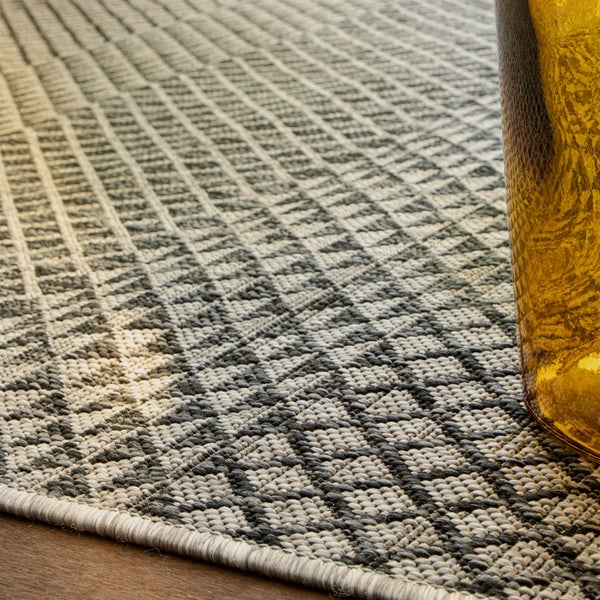 Tkaný koberec s šedo-bílým geometrickým kosočtvercovým vzorem a obšitým okrajem na dřevěné podlaze.