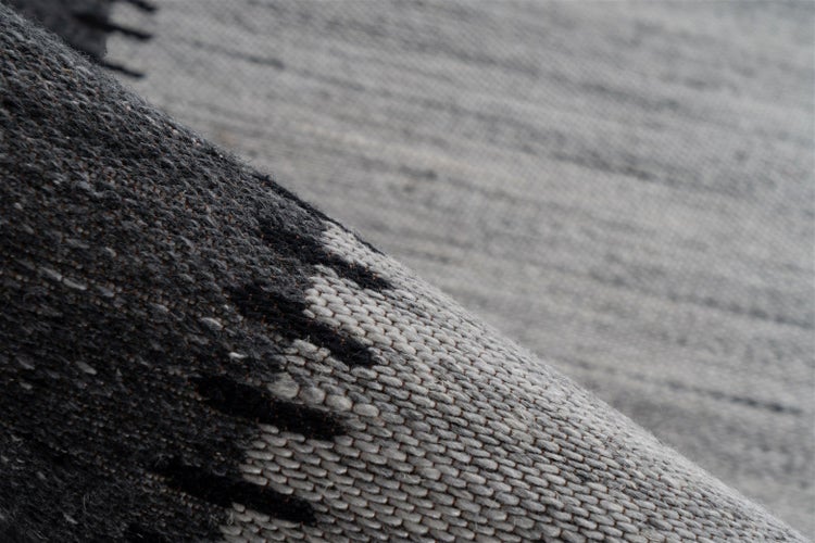 Detailni pohled na tkany koberec s sedym a tmave sedym geometrickym vzorem a hrubou texturou.