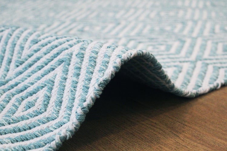 Detail tkaného koberce s geometrickým vzorem v bledě modré a bílé barvě, ohrnutý okraj na dřevěné podlaze.