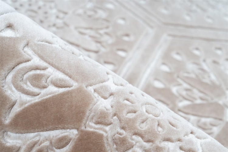 Detailní záběr na béžový koberec s reliéfním ornamentálním vzorem a měkkou strukturou s vysokým a nízkým vlasem.