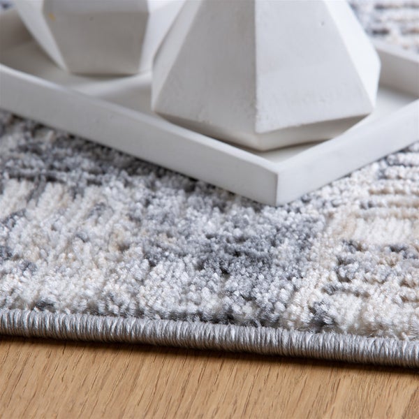 Detailní záběr na vzorovaný koberec v šedé a bílé barvě s obšitým okrajem na dřevěné podlaze.