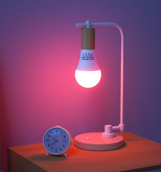 Moderní stolní lampa s chytrou LED žárovkou a analogovým budíkem na dřevěném stole.