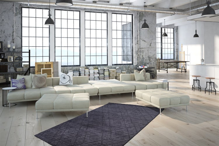 Prostorný a světlý loft v industriálním stylu s vysokými okny, béžovou sedací soupravou, dřevěnou podlahou a tmavým kobercem v obývacím pokoji.