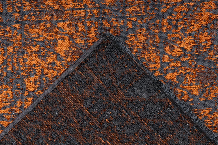 Detailní záběr na tkaný koberec v oranžové a tmavě šedé barvě s vintage vzorem, zobrazující strukturu povrchu a zahnutý roh.