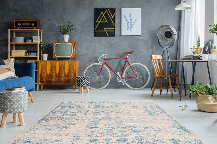 Obývací pokoj ve vintage stylu s kobercem s orientálním vzorem, modrou pohovkou, dřevěnou komodou, kolem u šedé stěny a pokojovými rostlinami.