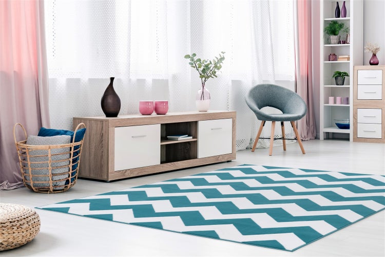 Světlý obývací pokoj s TV stolkem, šedým křeslem a kobercem se cikcak vzorem na podlaze.