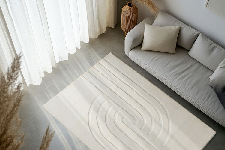 Obývací pokoj s moderním kobercem s geometrickým reliéfním vzorem na betonové podlaze, vedle pohovky a závěsů ve světlém interiéru.