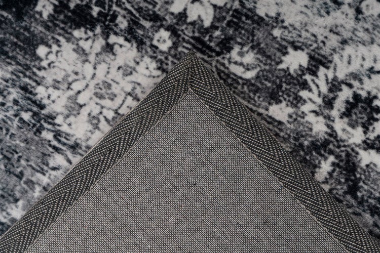 Detailní záběr na roh šedo-bílého koberce se vzorovaným vlasem ve vintage stylu a tkanou zadní stranou se zesíleným okrajem.