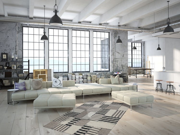 Velký otevřený loft v industriálním stylu s obývacím pokojem, pohovkou, geometrickým kobercem, vysokými okny a stěnami z neomítnutého betonu.