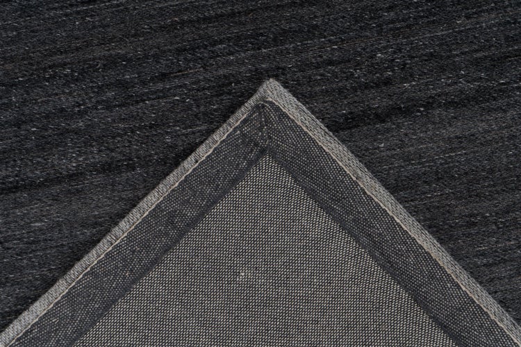 Detailní záběr tmavého texturovaného koberce se zaměřením na tkanou zadní stranu a roh.