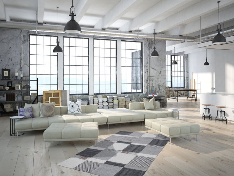 Prostorný loft v industriálním stylu s otevřenou kuchyní, velkou pohovkou, vysokými okny, betonovými stěnami a dřevěnou podlahou.