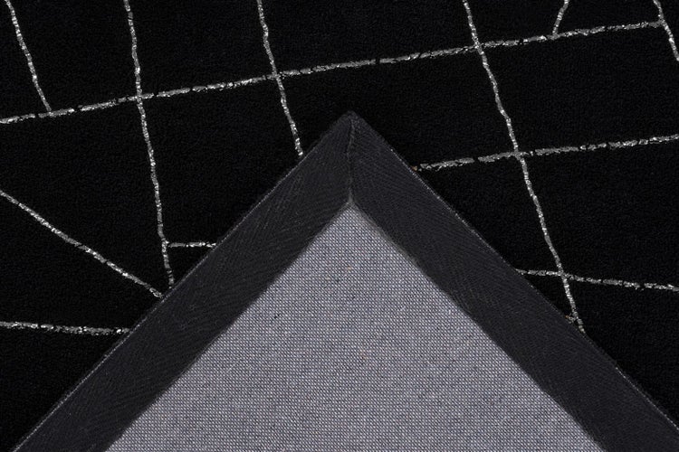Černý koberec se stříbrným geometrickým vzorem, detailní pohled na spodní stranu v ohrnutém rohu.