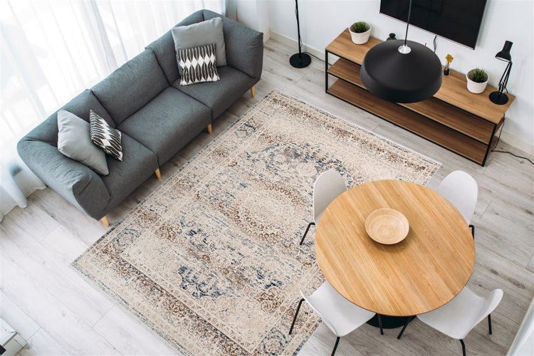 Obývací pokoj se šedou pohovkou, kulatým dřevěným stolem a kobercem s orientálním vzorem. Světlý moderní design s dřevěnými a kovovými prvky.