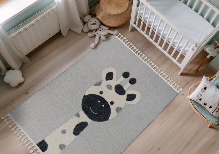 Dětský koberec s motivem žirafy a třásněmi v útulném dětském pokoji s postýlkou na dřevěné podlaze.