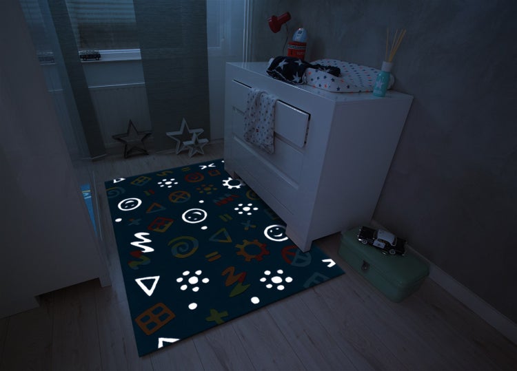 Dětský pokoj se svítícím kobercem s geometrickými tvary a emotikony před bílou komodou v tmavé atmosféře.