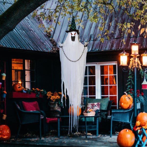 Dekorativní halloweenská scéna se závěsným duchem s čarodějnickým kloboukem a řetězy před domem s dýněmi a podzimní výzdobou.